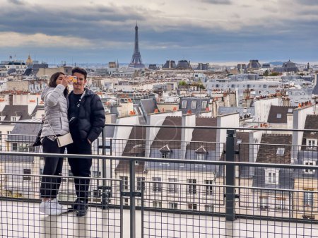 Téléchargez les photos : Paris France - 04.06.2022 : Sur le toit de Paris, un jeune couple prend un selfie avec téléphone portable avec une belle vue générale sur la ville et la tour eiffel en arrière-plan. Ciel nuageux avec copie - en image libre de droit