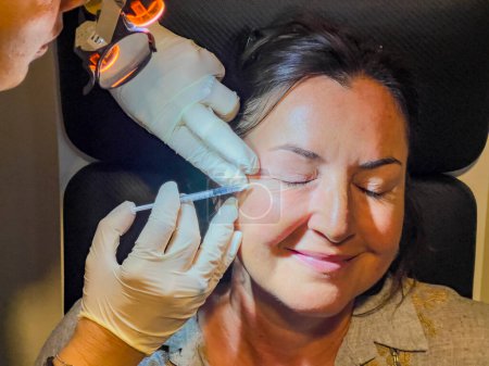 Happy smiling woman receiving facial aesthetic rejuvenation and Botox injections in the eye area in a beauty clinic. Anti-aging concept background, copy space. High angle view.