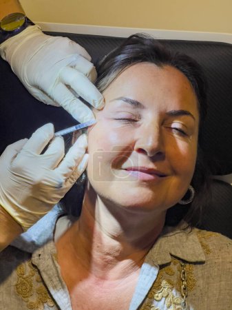 Smiling brunette woman, eyes closed, receiving facial aesthetic rejuvenation and botox injections in a beauty clinic. Anti-aging concept background, copy space. High angle view.
