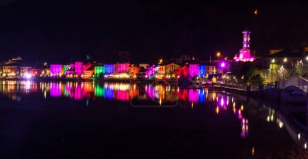 Photo for Porto Ceresio, Italy: 12-25-2021: Houses illuminated by searchlights on Christmas night, by colored lights reflecting on the water of the Lake of Lugano - Royalty Free Image