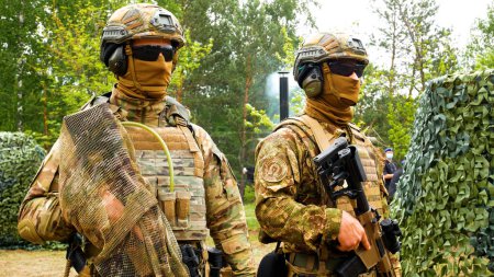 Foto de Kharkiv, Ucrania - 29 de mayo de 2021: Dos soldados en uniformes militares, chalecos antibalas, cascos, gafas oscuras con armas están cerca de una red de camuflaje de equipo militar. Guerra ruso-ucraniana 2022-2023 - Imagen libre de derechos