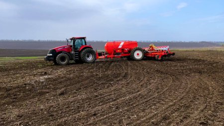 Foto de Kharkiv, Ucrania - 19 de abril de 2023: Un tractor con una sembradora recorre un campo en primavera y siembra trigo. Campaña de siembra para agricultores. Hay un campo verde en el fondo - Imagen libre de derechos