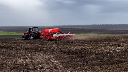 Foto de Kharkiv, Ucrania - 17 de abril de 2023: Un tractor con una sembradora atraviesa un campo en primavera y siembra grano de trigo en el suelo. Cultivo de cereales. agricultores ucranianos están sembrando grano para el - Imagen libre de derechos
