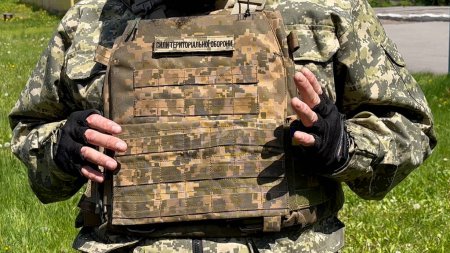 Téléchargez les photos : Un soldat ukrainien en uniforme de camouflage et en gilet pare-balles portant l'inscription des Forces de défense territoriale se tient debout et tient la main sur un gilet pare-balles, des discussions, des haussements d'épaules. Dans le - en image libre de droit