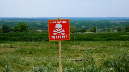 A warning sign with the inscription Caution, mines, with the image of a skull against the background of a Ukrainian village. Mined areas after the de-occupation of Ukrainian villages during the