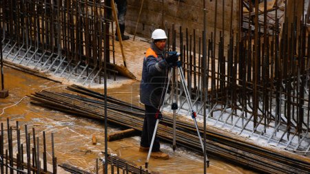 Foto de Kharkiv, Ucrania - 04 de diciembre de 2023: Un constructor se encuentra en una obra de construcción sobre una base cementada entre el refuerzo de metal, mira un dispositivo de medición y establece el nivel. Construcción de la - Imagen libre de derechos