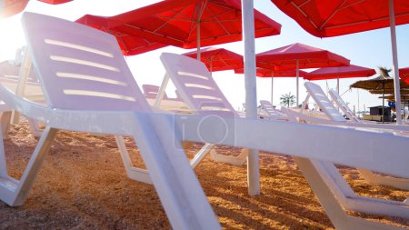Empty white plastic sun loungers on sandy beach under red umbrellas. Summer holidays.