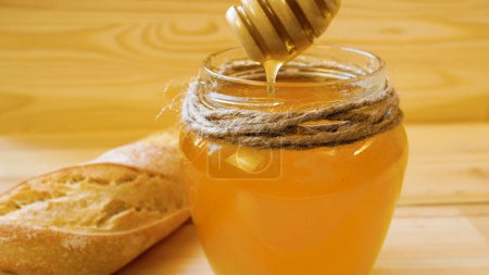 Liquid honey drips from a wooden spoon into a jar. A grain baguette lies nearby. Honey and bee products in the diet.