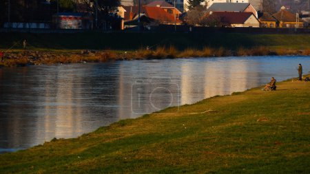 A small river in the setting sun. Fishermen are sitting on the green shore. In the background are residential buildings. Concept of quiet life by the river, rest on the river bank.
