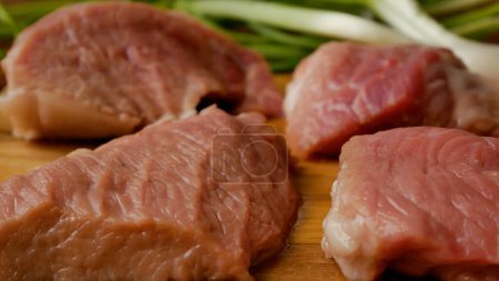 Stücke von frischem Schweinefleisch in Scheiben auf einem Schneidebrett. Im Hintergrund sind grüne Zwiebeln zu sehen. Fleischgerichte in der Küche.