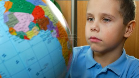 Un garçon de 6-7 ans tourne et examine un globe. Enseigner aux enfants à l'école. Enfants et géographie.