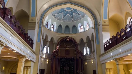 The synagogue building is from the inside. A two-storey hall with a domed ceiling. The suns rays enter through many windows. A religious building for Jewish prayers.
