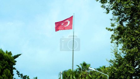 Flag of Turkey on a tall flagpole against blue sky. There are green trees on the sides.