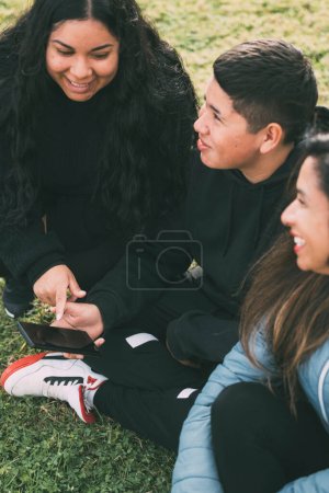 Foto de Three individuals of Hispanic-Latino ethnicity sitting on the lush green grass of a park, with smart device. Relaxed and smiling expressions on their faces they are enjoying a moment of leisure amidst - Imagen libre de derechos