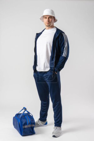 Foto de Un hombre en chándal, camiseta blanca y panama se encuentra cerca de una gran bolsa deportiva azul - Imagen libre de derechos