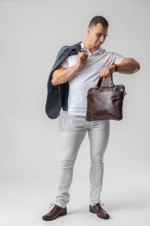 Foto de Un hombre con ropa casual mira su reloj y sostiene un maletín de cuero y una chaqueta en sus manos. Modelo posando sobre fondo blanco - Imagen libre de derechos