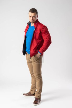 Foto de Un hombre atlético con una chaqueta roja, una camiseta azul y pantalones beige se para y sostiene sus manos en los bolsillos. Modelo posando sobre un fondo blanco. - Imagen libre de derechos