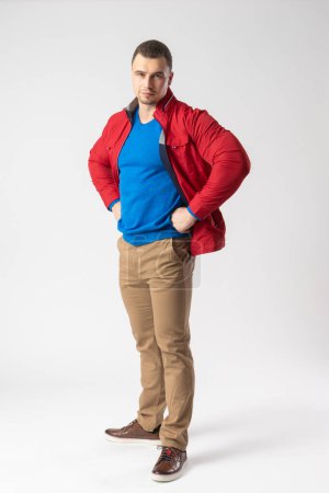 Foto de Un hombre de constitución atlética en una chaqueta roja, camiseta azul y pantalones beige está de pie akimbo. Modelo posando sobre un fondo blanco. - Imagen libre de derechos