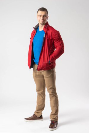 Foto de Un hombre atlético con una chaqueta roja, una camiseta azul y pantalones beige se para y sostiene sus manos en los bolsillos. Modelo posando sobre un fondo blanco. - Imagen libre de derechos