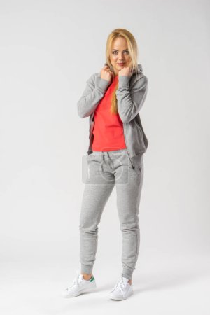 Photo for A young blonde woman in a gray tracksuit, red tank top and white sneakers poses in the studio on a white background. - Royalty Free Image