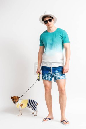 Foto de Joven hombre moreno con un sombrero blanco, gafas de sol, traje de playa y chanclas con un perro Jack Russell Terrier con una correa posando en el estudio sobre un fondo blanco - Imagen libre de derechos