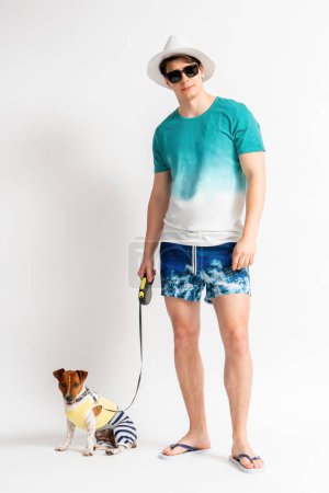 Foto de Joven hombre moreno con un sombrero blanco, gafas de sol, traje de playa y chanclas con un perro Jack Russell Terrier con una correa posando en el estudio sobre un fondo blanco - Imagen libre de derechos