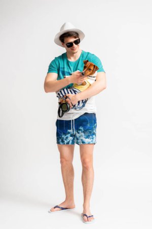 Foto de Joven hombre moreno con un sombrero blanco, gafas de sol, traje de playa y chanclas con un perro Jack Russell Terrier en sus brazos posando en el estudio sobre un fondo blanco - Imagen libre de derechos