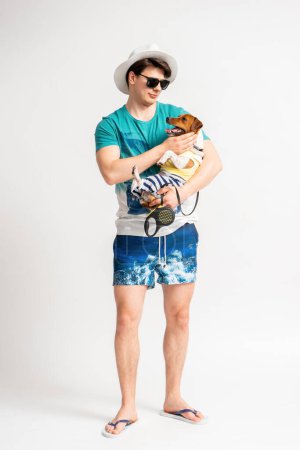 Foto de Joven hombre moreno con un sombrero blanco, gafas de sol, traje de playa y chanclas con un perro Jack Russell Terrier en sus brazos posando en el estudio sobre un fondo blanco - Imagen libre de derechos