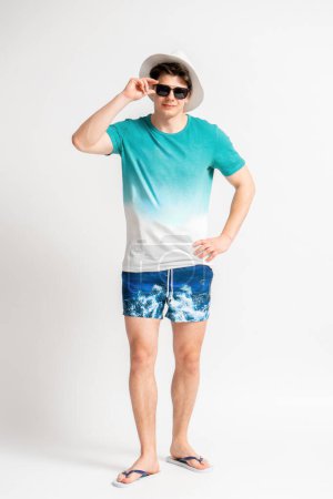 Foto de Joven hombre moreno con un sombrero blanco, traje de playa y chanclas ajustando sus gafas de sol posando en el estudio sobre un fondo blanco - Imagen libre de derechos