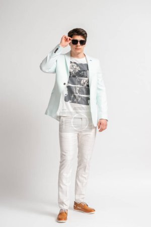 Foto de Joven hombre de cabello oscuro con una chaqueta casual de color claro y camiseta ajustando sus gafas de sol mientras posa de cuerpo entero en el estudio sobre un fondo blanco - Imagen libre de derechos