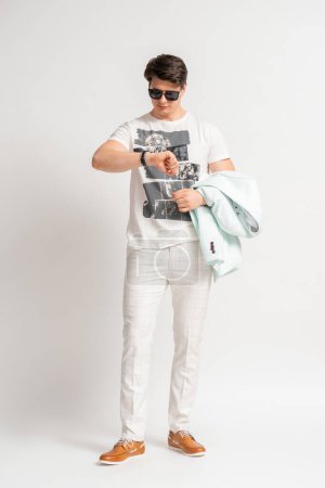 Foto de Joven hombre moreno con ropa casual de verano mira su reloj y sostiene una chaqueta en sus manos posando en el estudio sobre un fondo blanco - Imagen libre de derechos