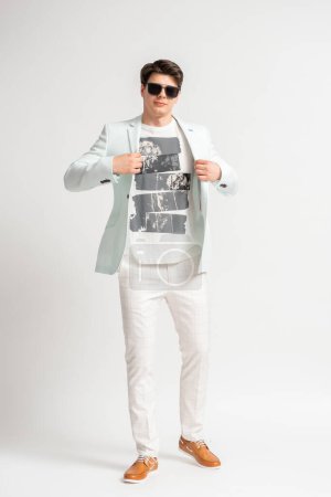 Foto de Joven hombre moreno con una chaqueta casual de color claro y gafas de sol posando en el estudio sobre un fondo blanco - Imagen libre de derechos