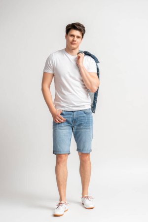 Foto de Hombre joven de cabello oscuro con una camiseta blanca casual de verano, pantalones cortos de mezclilla y una chaqueta colgada sobre su hombro, posando en el estudio sobre un fondo blanco - Imagen libre de derechos