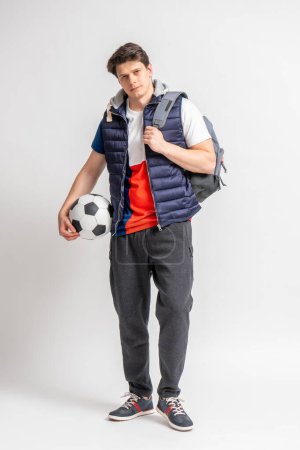 Foto de Joven hombre moreno en ropa deportiva casual con una pelota de fútbol y una mochila posando en toda su longitud sobre un fondo blanco - Imagen libre de derechos