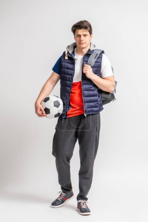 Foto de Joven hombre moreno en ropa deportiva casual con una pelota de fútbol y una mochila posando en toda su longitud sobre un fondo blanco - Imagen libre de derechos