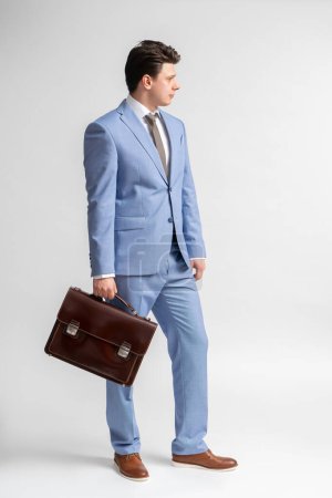 Foto de Un joven moreno con un traje de negocios azul, camisa blanca y corbata con un maletín de cuero en las manos posa en pleno crecimiento en el estudio sobre un fondo blanco - Imagen libre de derechos