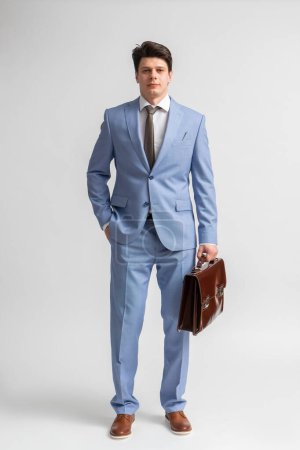 Foto de Un joven moreno con un traje de negocios azul, camisa blanca y corbata con un maletín de cuero en las manos posa en pleno crecimiento en el estudio sobre un fondo blanco - Imagen libre de derechos