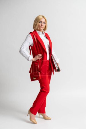 Photo for Young blonde business woman in a red casual trouser suit with a large office folder and a women's handbag on her shoulder posing on a white background - Royalty Free Image