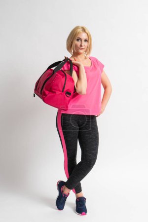 Photo for A young blonde woman in a pink tank top, sports leggings and sneakers holds a large fitness training bag in her hands. Model posing in the studio on a white background. - Royalty Free Image
