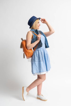 Photo for Happy young blonde woman in a blue casual summer dress, a denim vest with a backpack straightens her hat and poses in the studio on a white background. - Royalty Free Image