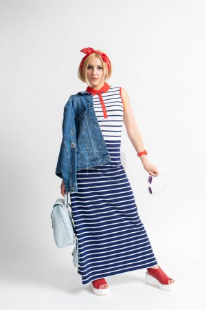 Photo for Young blonde woman in a long casual striped dress with a red bandana on her head, a backpack, sunglasses and a denim jacket in her hand posing on a white background - Royalty Free Image