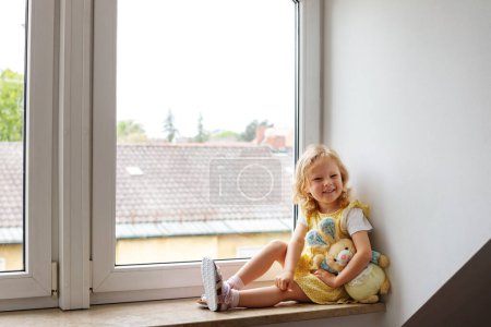 Foto de Lindo bebé en casa en una habitación blanca se sienta cerca de la ventana. Hermosa niña con oso de peluche. Un niño con el pelo rizado - Imagen libre de derechos