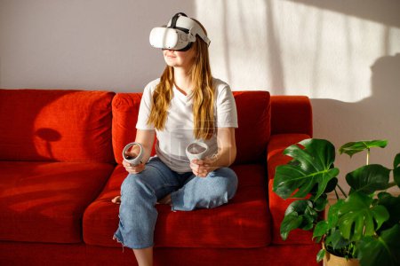 Frau plus Größe in virtueller Realität durch VR-Brille auf Sofa mit lustigen und fröhlichen Momenten im heimischen Wohnzimmer beobachtet