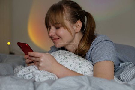 woman at home in bed with smartphone. Nice App. Smiling Woman Using Smartphone In Bed, Happy Female Relaxing With Mobile Phone In Bedroom, Browsing New Application, Copy Space