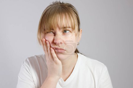 Jeune femme misérable éprouvant des maux de dents sévères, pressant la paume sur la joue, isolée sur fond gris