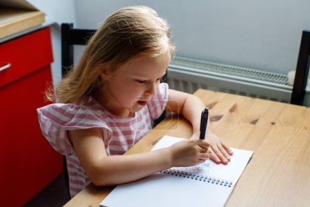 a girl sits at the table in the kitchen and draws in a notebook. home education of a child