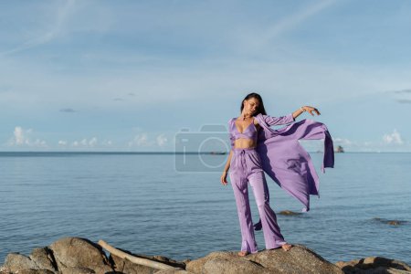 Téléchargez les photos : Jeune belle femme d'humeur romantique, vêtements en soie lilas, sur la plage sur fond de mer et pierres au coucher du soleil - en image libre de droit