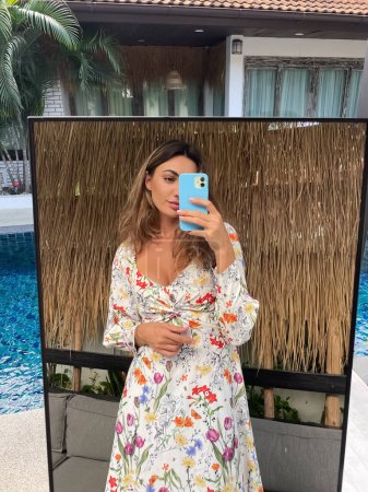 Téléchargez les photos : Femme en robe d'été avec fleur imprimé printemps à la maison en plein air prendre selfie photo sur le téléphone dans le miroir pour les médias sociaux, histoires, vertical - en image libre de droit