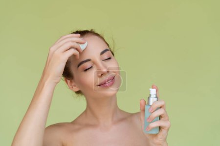 Beauty portrait of young topless red hair woman with bare shoulders on green background holds serum for youth and skin hydration and sponge