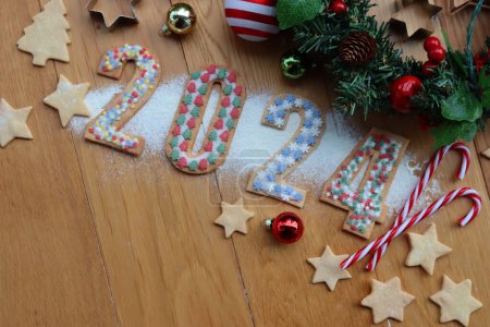 Foto de Galletas de corteza corta con espolvoreos de colores en forma de números 2024 con guirnaldas de pino y adornos en mesa de madera - Imagen libre de derechos
